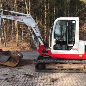 May include: A white and red Takeuchi TB145 mini excavator with a bucket attachment. The excavator is on a paved surface with a forest background. The machine has a cab with windows and a boom arm.