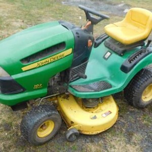 Peut inclure: Tondeuse autoportée John Deere verte et jaune. La tondeuse a un capot vert avec le logo John Deere et un siège jaune. Elle possède un plateau de coupe jaune et des pneus noirs avec des jantes jaunes.
