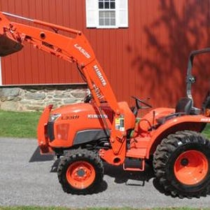 Pode incluir: Um trator laranja Kubota L3301 com carregador frontal e barra de proteção. O trator tem pneus pretos e está estacionado em uma superfície pavimentada em frente a um celeiro vermelho. Um trator verde é visível ao fundo.