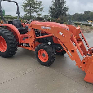 Op de afbeelding: Een oranje Kubota-tractor voorzien van een voorlader met bak, een zwarte bestuurdersstoel, een rolbeugel ter bescherming en grote zwarte rubberen banden met oranje velgen, geplaatst op een verhard oppervlak.