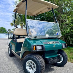 Op de afbeelding: Een groene Club Car golfkar met een beige dak en stoelen. De kar heeft een voorruit, koplampen en zwarte banden met witte velgen. De kar staat geparkeerd op een verharde ondergrond met bomen op de achtergrond.