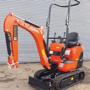 May include: An orange Kubota mini excavator with a black seat and tracks. The excavator has a black arm and bucket, with the "Ardent" logo visible. The machine is on a concrete surface in front of a corrugated metal wall.