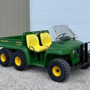 May include: A green John Deere Gator 6x4 utility vehicle with yellow seats and black tyres. The vehicle has a windscreen and a cargo bed. The tyres have yellow rims. The vehicle is parked on gravel.