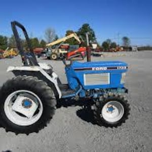 Può includere: Un trattore Ford 1700 blu e bianco con pneumatici neri è parcheggiato su una superficie di ghiaia. Il trattore ha un roll bar ed è circondato da altri veicoli da costruzione sotto un cielo azzurro.