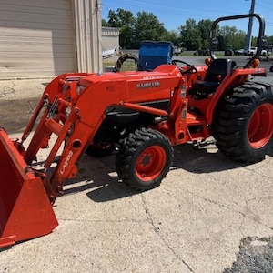 Pode incluir: Um trator Kubota laranja com carregador frontal e um acessório de corte traseiro. O trator tem pneus pretos e um assento preto. O número do modelo é visível na lateral do trator.