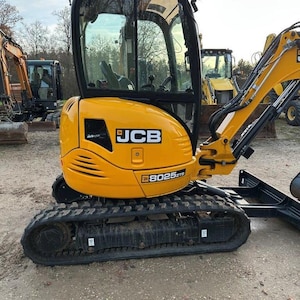 Puede incluir: Mini excavadora JCB 8025 ZTS amarilla con cabina y orugas negras. La excavadora está estacionada al aire libre, con otros equipos de construcción visibles en el fondo. El logotipo de JCB se muestra de forma destacada.