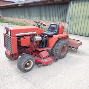 May include: A red Wheel Horse tractor with a black seat, steering wheel, and large tyres. The tractor has a front grill, headlights, and a rear attachment. The tractor is parked on a concrete surface.