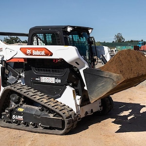 May include: A white and orange Bobcat T66 compact track loader with a full bucket of brown material. The loader is parked on a dirt surface next to a truck. The Bobcat logo is visible on the side.