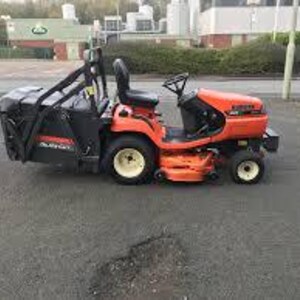 Pode incluir: Um cortador de relva a gasolina laranja e preto com um recipiente de recolha preto preso na parte traseira. O cortador de relva tem um assento, volante e rodas amarelas. O cortador de relva está estacionado numa superfície pavimentada.