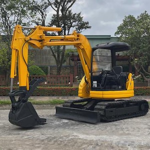 Op de afbeelding: Een gele graafmachine met een zwarte bak en rupsbanden. Het bouwmachine heeft een zwarte cabine en staat op een betonnen ondergrond. De graafmachine staat voor een groene en bruine achtergrond.