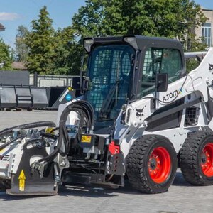 Könnte beinhalten: Ein weiß-schwarzer Bobcat S590 Kompaktlader mit einem frontmontierten Fräsvorsatz. Der Lader hat schwarze Reifen mit roten Felgen und eine verglaste Kabine. Die Maschine steht an einem sonnigen Tag im Freien.