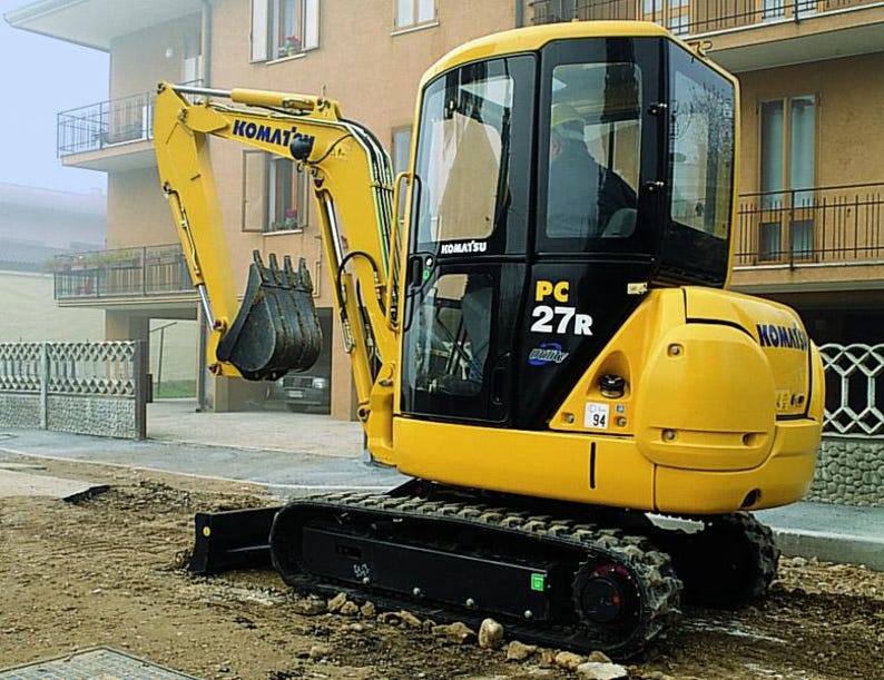 May include: A yellow Komatsu PC27R mini excavator with a black cab and tracks. The excavator has a bucket attachment and is parked on a construction site. The Komatsu logo is visible on the arm and body of the machine.