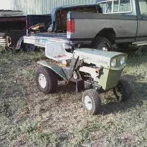 May include: An olive green riding lawn mower with a white seat and black tires is parked on a grassy area. A gray pickup truck is in the background. The mower has two headlights and a front bumper.