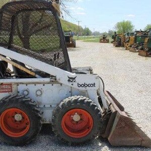 May include: A white Bobcat skid-steer loader with orange wheels and a front bucket. The loader has a black mesh safety cage and the Bobcat logo. Other construction equipment is visible in the background.