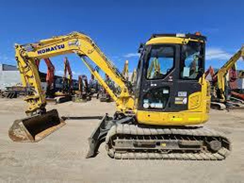 May include: A yellow Komatsu excavator with a black cab and a metal bucket. The excavator is on a gravel surface with other construction equipment in the background. The sky is blue.