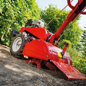 May include: A red and black garden tiller with a white tire is angled on a dirt slope. The tiller has a blade attachment and a handle. Green foliage and trees are in the background.