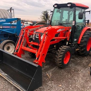 Può includere: Un trattore rosso brillante con un caricatore frontale nero e una benna nera. Il trattore ha pneumatici grandi e neri con cerchi rossi e una cabina di vetro. Un trattore Ford blu è visibile sullo sfondo.