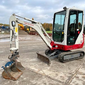 Puede incluir: Una excavadora compacta Takeuchi, con una cabina blanca y roja y un cerramiento de vidrio. La excavadora tiene un brazo y un cucharón grises y está situada sobre una superficie de hormigón. La máquina está diseñada para tareas de construcción y movimiento de tierras.