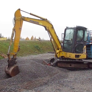 Op de afbeelding: Een gele Komatsu-graafmachine met een zwarte cabine staat bovenop een hoop grind. De arm van de graafmachine is uitgestrekt en de bak is open. Andere bouwmachines zijn zichtbaar op de achtergrond.