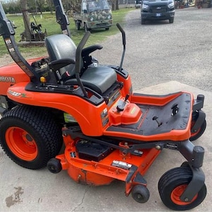 Op de afbeelding: Een oranje Kubota Z227 zero-turn grasmaaier. De maaier heeft een zwarte stoel, zwarte banden en oranje velgen. De maaier staat op een betonnen ondergrond.