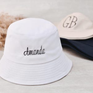 Puede incluir: Sombrero de pescador blanco bordado con el nombre "Amanda" en escritura negra. Se ven dos sombreros de pescador adicionales en el fondo, uno beige con "GB" y otro azul marino con "TH". Los sombreros son informales y elegantes.