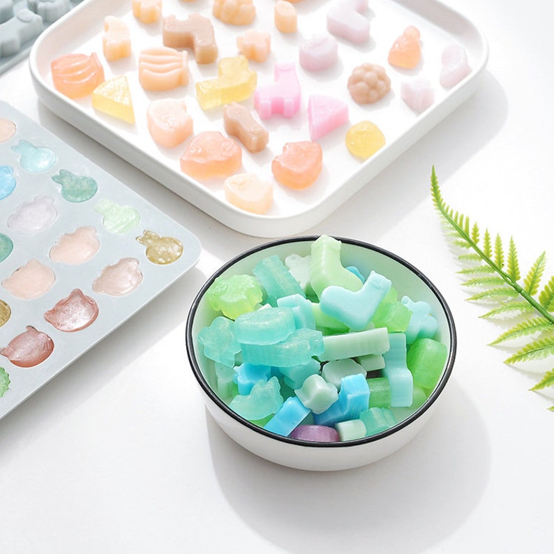 May include: A white bowl filled with small, colourful, handmade soap bars in various shapes, including stars, hearts, and rectangles. The soap bars are in shades of blue, green, and purple.