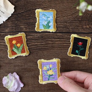 May include: Four embroidered iron-on patches in the shape of picture frames with different floral designs. The frames are gold with a white background. The flowers are yellow, white, red, and purple.