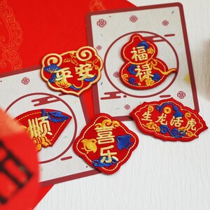 Könnte beinhalten: Eine Sammlung von bestickten Aufnähern in Rot, Gelb und Blau. Die Aufnäher zeigen chinesische Schriftzeichen und dekorative Designs. Sie sind auf roten und weißen Karten angeordnet, was auf ein kulturelles Thema hindeutet.