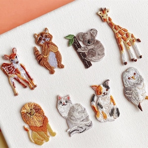 Könnte beinhalten: Set mit 10 bestickten Bügelbildern mit verschiedenen Tieren, darunter ein Reh, ein Waschbär, ein Koala, eine Giraffe, eine Eule, eine Katze und ein Löwe. Die Patches sind alle in verschiedenen Farben und Stilen.