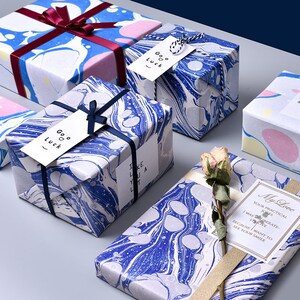 Puede incluir: Cuatro cajas de regalo envueltas en papel marmoleado azul y blanco con etiquetas de regalo blancas y negras que dicen "Goo Luck" y "I Like You A Lot". Una caja de regalo est&aacute; envuelta en papel marmoleado rosa, amarillo y azul. Una quinta caja de regalo est&aacute; envuelta en papel marmoleado azul y blanco con una cinta dorada y una etiqueta de regalo blanca que dice "My Love Your Practical Jones I Was IBerate- I Love Because I Want to See Your Smile"