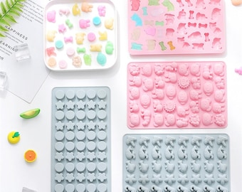 May include: Four silicone candy moulds with different designs. The moulds are on a white surface. The moulds are pink, blue, and grey. The designs include circles, stars, animals, and sea creatures.
