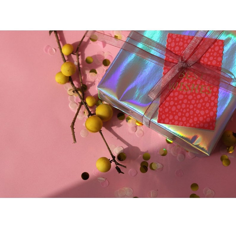 May include: A holographic gift box with a pink ribbon and a red card with white polka dots and the text "Best Wishes". The box is surrounded by gold confetti on a pink background.