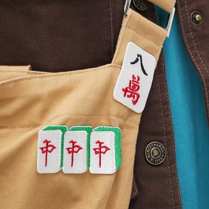 May include: A brown and blue denim jacket with a tan canvas bag attached. The bag has three green and white mahjong tiles with red Chinese characters that say "中" and a white patch with red Chinese characters that say "八萬".