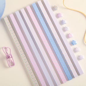 May include: A collection of ten long, narrow paper strips featuring various pastel patterns including diamonds, checks, dots, and stripes in shades of pink, blue, and yellow, accompanied by several small folded paper stars and a light pink clip.