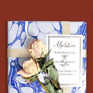 Puede incluir: Una caja de regalo blanca envuelta en papel marmoleado azul y blanco. La caja est&aacute; atada con una cinta dorada y tiene una etiqueta de regalo blanca con el texto "My Love Your Practical Jokes I Was Deliberately Move Cause I Want To Your Smile". Rosas secas est&aacute;n unidas a la caja.