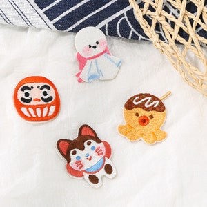 以下が含まれることがあります： 日本のデザインが施された4つの刺繍パッチ。ダルマ人形、てるてる坊主、ハスキー犬、たこ焼きのタコが含まれています。パッチはカラフルで、キュートな漫画風です。