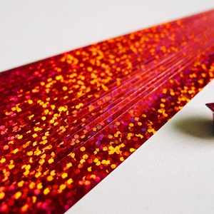 May include: A red iridescent paper strip with a red iridescent origami star. The paper strip has a glittery texture with small red, orange, and yellow dots.