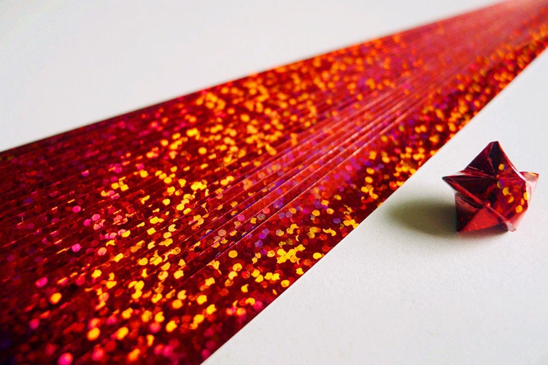 May include: A red iridescent paper strip with a red iridescent origami star. The paper strip has a glittery texture with small red, orange, and yellow dots.