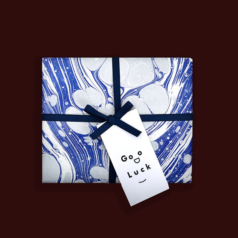 Puede incluir: Una caja de regalo blanca con un dise&ntilde;o de m&aacute;rmol azul y blanco. La caja est&aacute; atada con una cinta azul y tiene una etiqueta de regalo blanca que dice "Good Luck" en texto negro.
