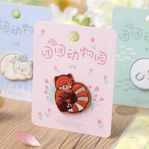 May include: A pink card with the text "团团动物园" in black lettering. The card features a red panda embroidered patch with brown accents. The text "浣熊" is printed below the patch. The card also has the text "Cardlover & Embroidery 信的恋人刺绣贴" printed at the bottom.