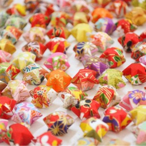 May include: A collection of colorful origami stars, folded from patterned paper. The stars are arranged in a random pattern on a white surface.
