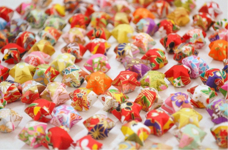 May include: A collection of colorful origami stars, folded from patterned paper. The stars are arranged in a random pattern on a white surface.