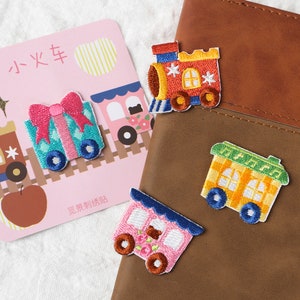 May include: Four embroidered iron-on patches featuring a train engine, a boxcar, a gift box, and a passenger car. The patches are colorful and feature a variety of details, including windows, wheels, and a bow on the gift box. The patches are on a brown leather background.