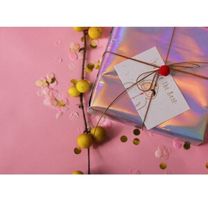 May include: A gift wrapped in iridescent holographic paper with a gold ribbon and a small red pom-pom. The gift tag is white with brown speckled paper and says "You're The Best". The gift is surrounded by pink and gold confetti on a pink background.