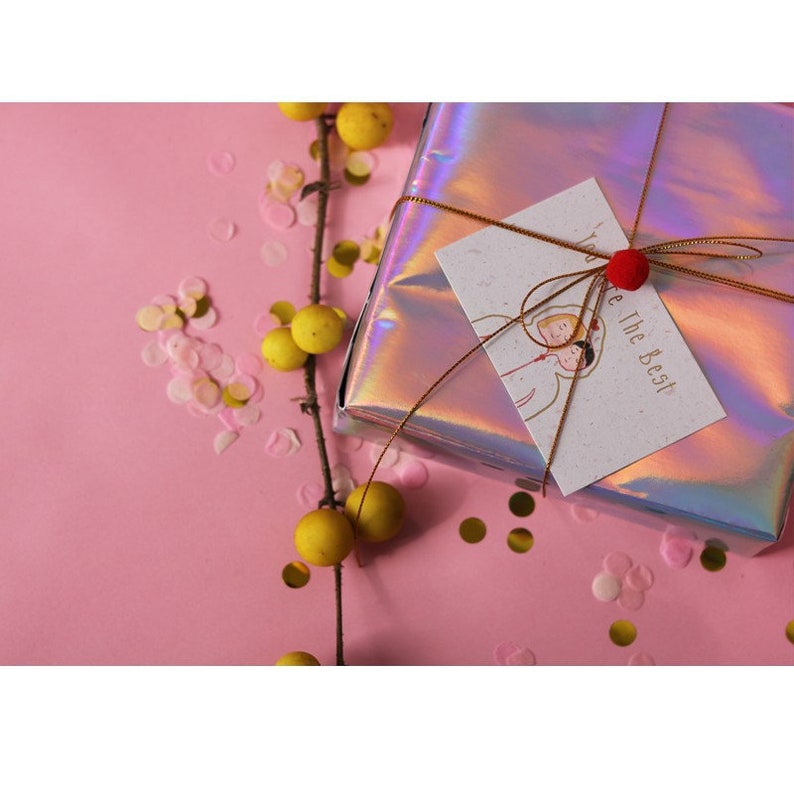 May include: A gift wrapped in iridescent holographic paper with a gold ribbon and a small red pom-pom. The gift tag is white with brown speckled paper and says "You're The Best". The gift is surrounded by pink and gold confetti on a pink background.