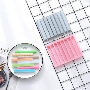 Puede incluir: Dos moldes de silicona para hacer crayones. Los moldes son grises y rosados y tienen ocho ranuras rectangulares. El molde rosado tiene la palabra "Crayons" impresa en cada ranura. También hay ocho crayones de colores en un plato blanco.