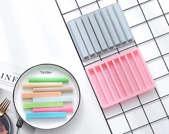 May include: Two silicone moulds for making crayons. The moulds are grey and pink and have eight rectangular slots. The pink mould has the word "Crayons" printed on each slot. There are also eight colourful crayons on a white plate.