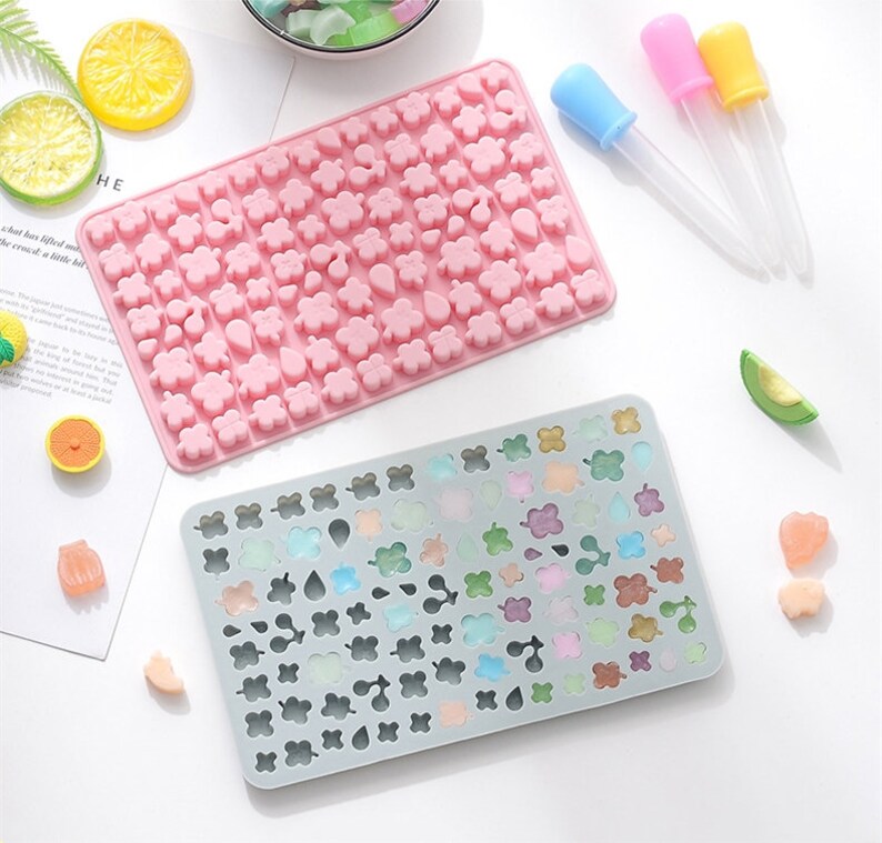 May include: Two silicone moulds for making candy or chocolate. The pink mould has a variety of small shapes, including flowers, hearts, and leaves. The grey mould has the same shapes but is filled with colourful candies.
