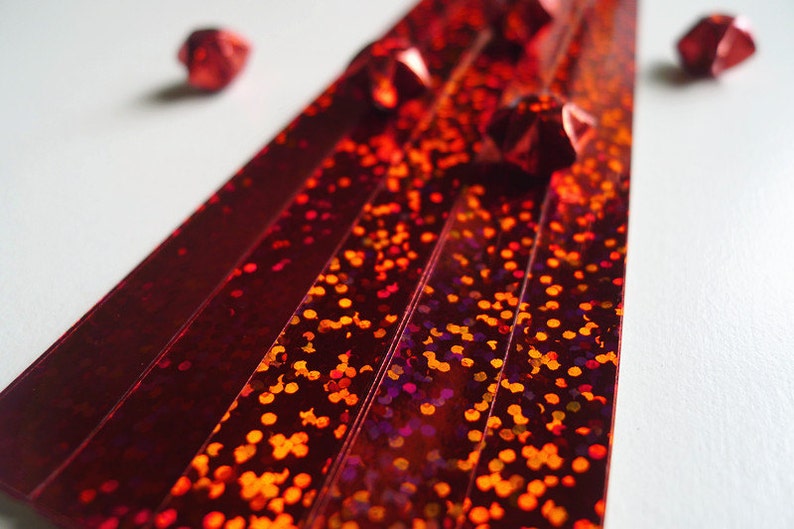 May include: Red iridescent paper strips with a glittery finish. The strips are arranged in a fan shape with red origami stars scattered around them.