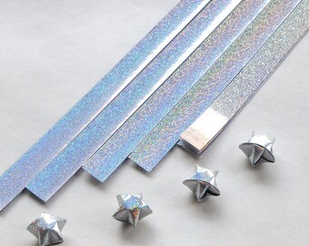 Puede incluir: Un conjunto de tiras de papel de origami plateado iridiscente con cuatro estrellas de origami dobladas. Las tiras de papel están dispuestas en diagonal.
